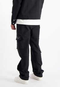 Zwarte cargo broek met meerdere zakken, gemaakt van textuurstof. Gecombineerd met een wit shirt, voorzien van een merkaccent op de zak.