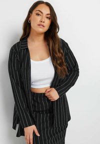 Blazer a righe bianche e nere con collo a rever, abbinato a un crop top bianco aderente. Il tessuto ha una consistenza morbida e una vestibilità comoda.