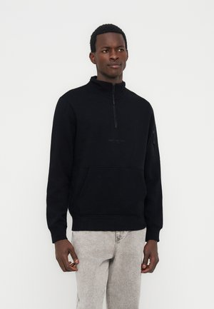 Mladi moški, oblečen v črno pulover s četrtinskim zadrgo in žepom spredaj ter svetlo sive kavbojke, stoji na belem ozadju.