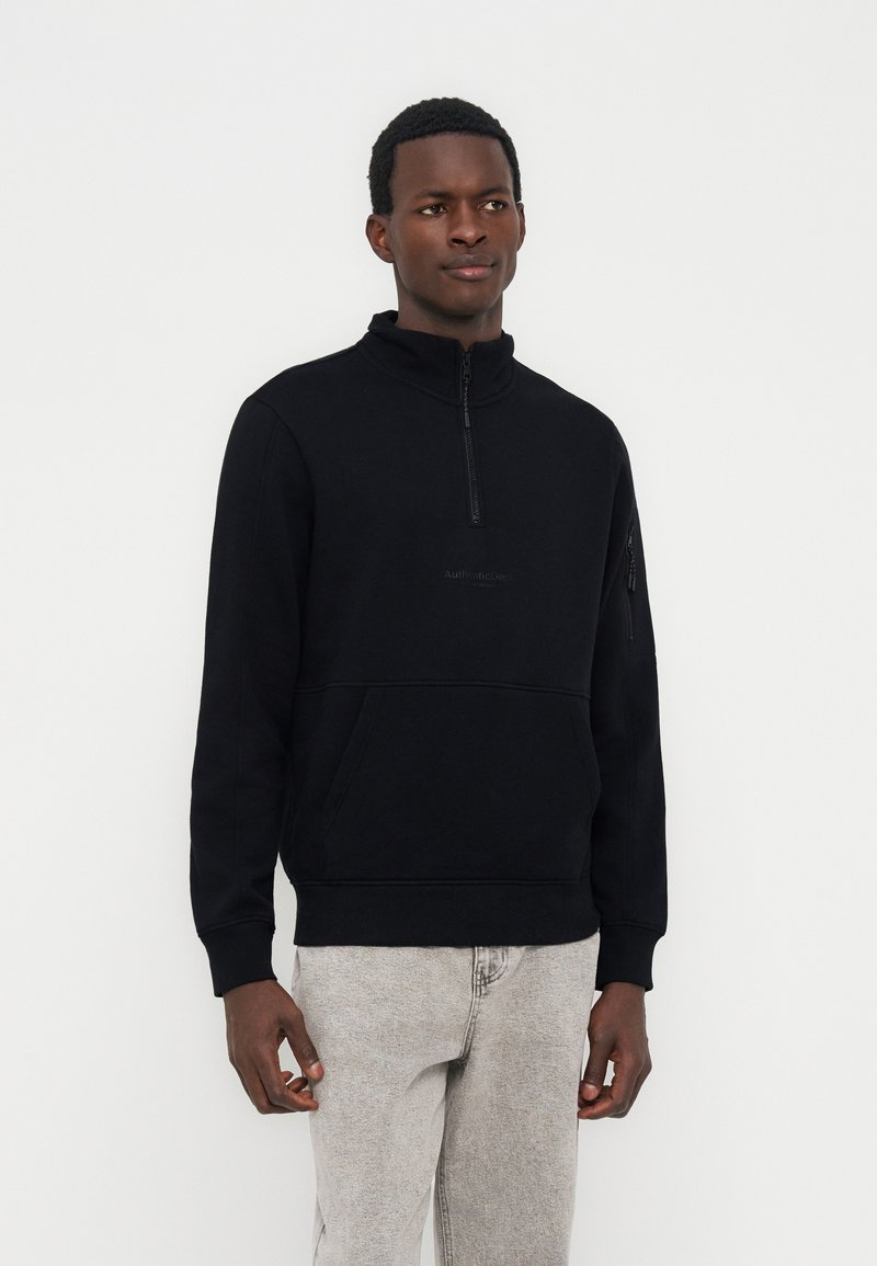 Νέος άνδρας που φοράει μαύρο φούτερ με φερμουάρ στο ένα τέταρτο και εμπρόσθια τσέπη, καθώς και ανοιχτό γκρι τζιν, στέκεται μπροστά σε άσπρο φόντο.