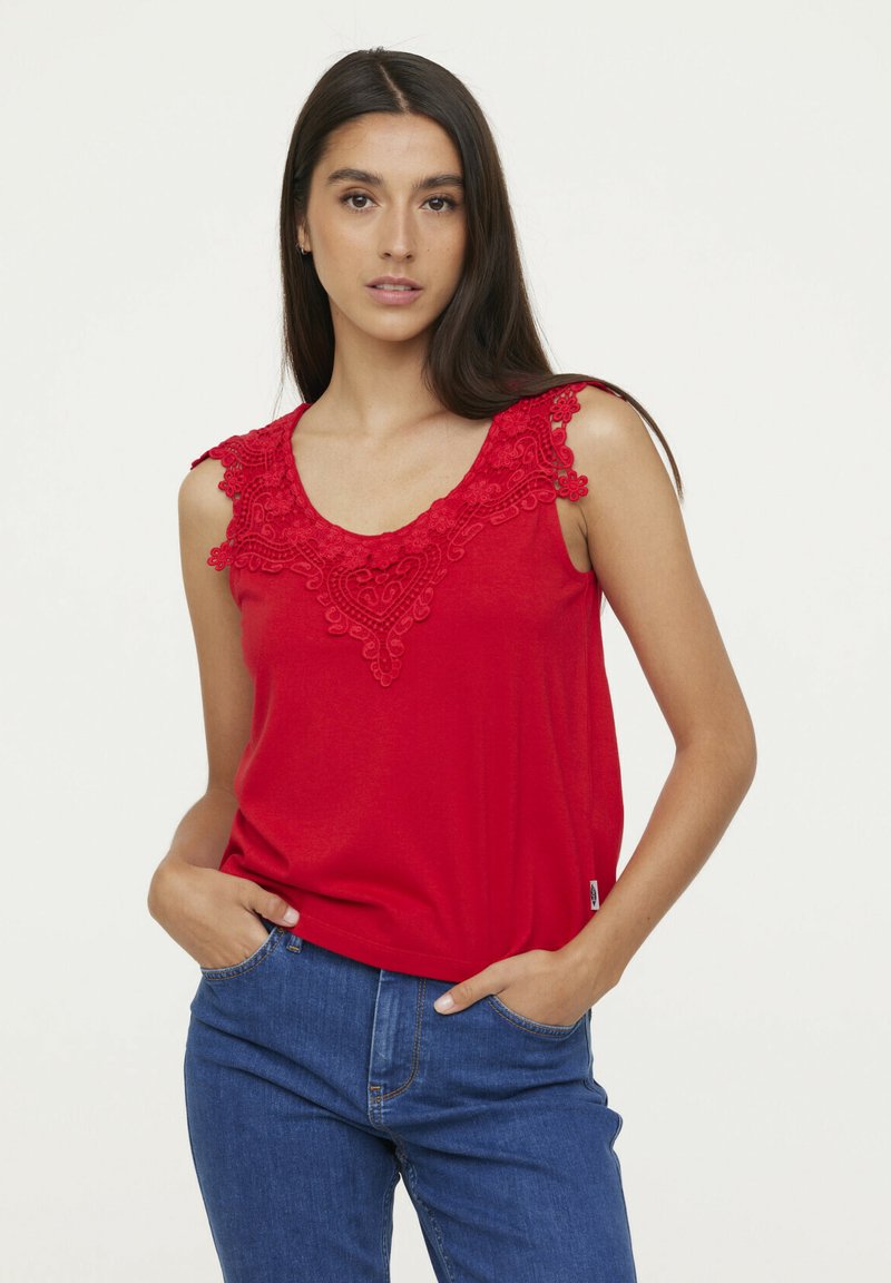 Débardeur rouge avec des détails en dentelle délicats autour de l'encolure, présentant une forme arrondie et une texture en coton douce. Porté avec un jean bleu.