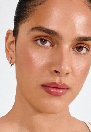 Vrouw met een gladde huid, bruine ogen, neutrale make-up, haar naar achteren getrokken, die een zilveren geometrische oorbel draagt aan het linkeroor tegen een effen achtergrond.