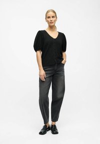 Kvinde i sort puffærmet knapbluse, falmede sorte højtaljede jeans og sorte loafers, der står med den ene hånd i lommen.