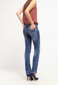 Kvinna i urtvättade blå jeans, en ärmlös vinröd topp och marinblå klackar, stående med händerna i bakfickorna.