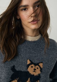 Grijze trui met een gebreid ontwerp van een hond in bruin en zwart. De trui heeft een geribde halslijn en een zachte, getextureerde afwerking.