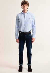 Camisa de botón azul claro, jeans de mezclilla oscura, cinturón de cuero marrón y zapatos de cordones marrones. El modelo está de pie de frente contra un fondo neutro.