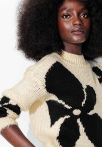 Pull en maille crème avec un grand motif floral noir, doté d'un col montant et de larges poignets. Tissu texturé avec un aspect douillet.