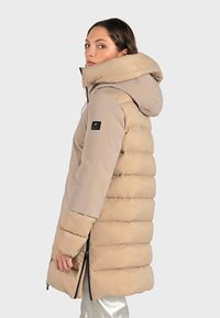 Beige gewatteerde jas met een capuchon, gestikt ontwerp en zijritsen. Kenmerkt zich door een merklint op de linkermouw. Glad, licht glanzend textuur.