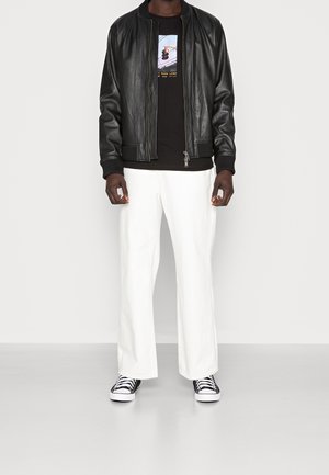 Mand iført sort læderjakke, sort grafisk t-shirt, hvide bukser og sort-hvide sneakers foran ensfarvet baggrund.