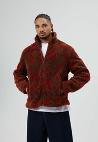 Veste en polaire duveteuse rouge et marron avec un col montant, une fermeture éclair et un motif texturé. Portée sur une chemise blanche et associée à un jean foncé.