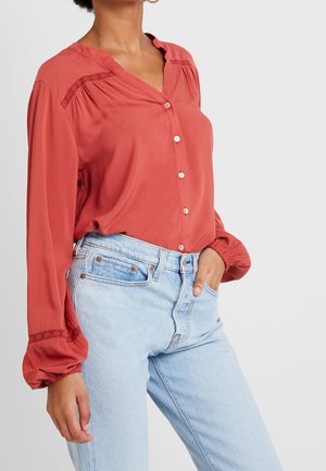 Kobieta ubrana w rdzawą, zapinaną na guziki bluzkę z koronkowymi detalami na rękawach oraz jasnoniebieskie, wysokowciskające jeansy, ręka w kieszeni, na szarym tle.