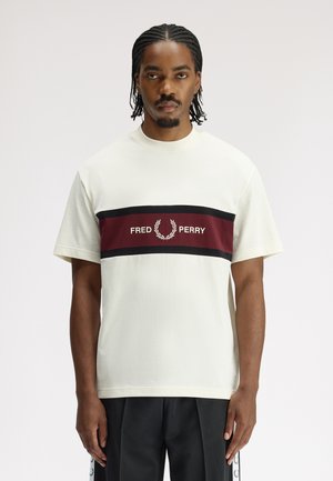 Homme aux cheveux tressés portant un T-shirt blanc avec une bande bordeaux et noire traversant la poitrine et le logo "Fred Perry", debout devant un fond blanc.