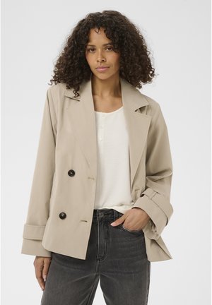 Femme aux cheveux bouclés portant une veste beige croisée, un haut blanc et un jean noir, debout avec une main dans la poche.