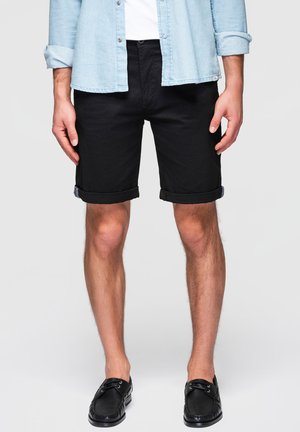 Uomo che indossa pantaloncini neri risvoltati, scarpe nere slip-on, t-shirt bianca e camicia azzurro chiaro a maniche lunghe arrotolate, in piedi davanti a uno sfondo semplice.