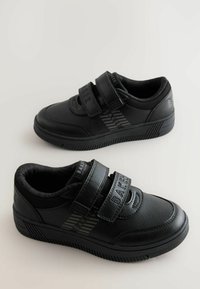 Schwarze Turnschuhe mit einer strukturierten Synthetik-Oberfläche, zwei Klettverschlüssen und einer gerippten Gummisohle mit dezentem Branding und gestreiften Akzenten.