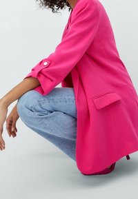 Persona agachada, vistiendo un blazer rosa brillante con las mangas arremangadas, jeans azul claro y zapatos rosa a juego contra un fondo claro y liso.