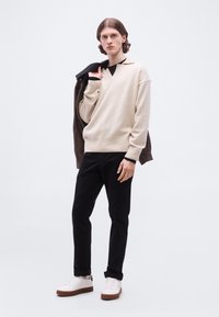 Νέος άνδρας με μακριά μαλλιά μέχρι τους ώμους φοράει μπεζ πουλόβερ, μαύρο παντελόνι, λευκά αθλητικά παπούτσια και κρατά ένα σκούρο μπουφάν περασμένο πάνω από τον έναν ώμο.