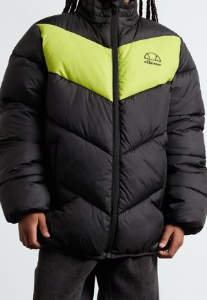 Czarna kurtka puchowa z neonowożółtym wzorem w kształcie szewrona na klatce piersiowej, zapinana na zamek z przodu, noszona przez osobę w ciemnych spodniach i z warkoczami.