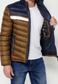Homme portant une veste matelassée bleu marine et marron, ouverte pour montrer la doublure bleu marine et l'étiquette, un t-shirt blanc et un jean bleu.