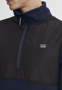 Pullover navy con colletto alto, caratterizzato da una chiusura a mezzo zip e una combinazione di pile e tessuto liscio. Include un patch con il logo.