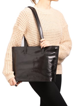 Woman in beige knit sweater and black pants holding large black leather tote bag with two shoulder straps.
