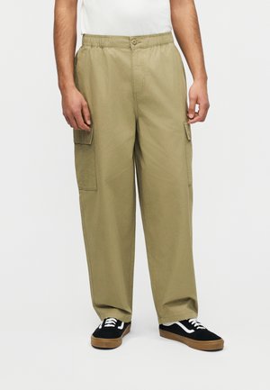 Man wearing loose-fitting khaki cargo pants with button closure and black sneakers with white laces, standing against a white background.