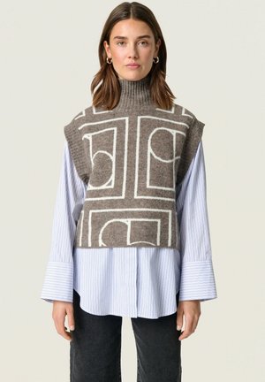 Mouwloze, bruine wollen sweater met witte geometrische patronen, gelaagd over een blauw-wit gestreept shirt met brede manchetten en donkere broek.