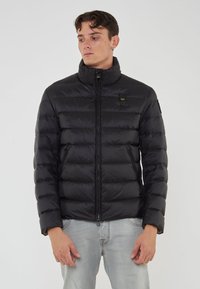 Giovane uomo che indossa un giubbotto trapuntato nero con colletto alto e tasche con zip, abbinato a jeans azzurri chiari, in piedi davanti a uno sfondo semplice.