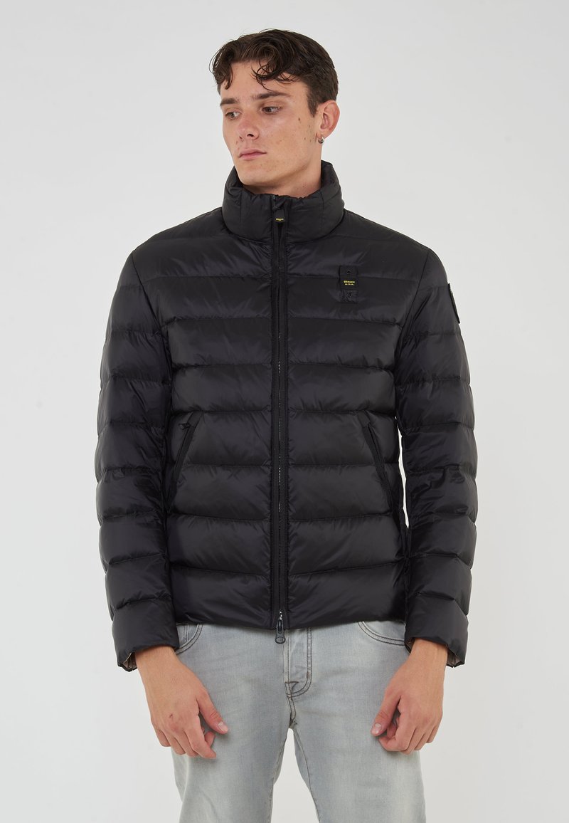 Giovane uomo che indossa un giubbotto trapuntato nero con colletto alto e tasche con zip, abbinato a jeans azzurri chiari, in piedi davanti a uno sfondo semplice.
