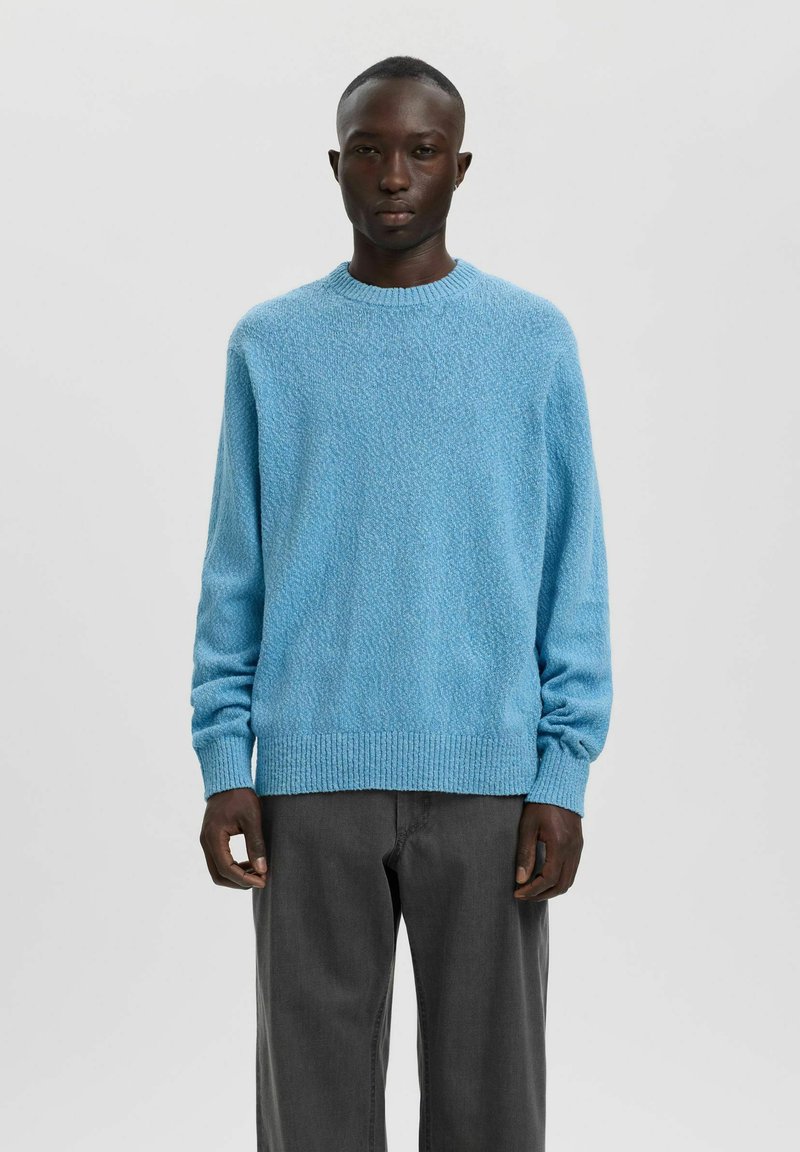 Homme portant un pull à manches longues bleu clair texturé et un pantalon gris foncé, debout devant un fond uni gris clair.