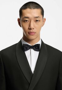 Tuxedo nero con risvolti in satin e camicia bianca. Presenta un papillon nero e una texture liscia. Capelli rasati.