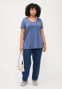 Una persona con cabello rizado lleva una camiseta azul con escote en V y unos jeans azules holgados, sosteniendo un pequeño bolso beige.