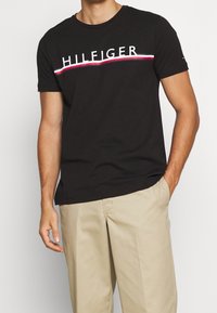 Camiseta de algodón negra con el logo blanco "HILFIGER" y una franja curva roja y blanca debajo. Diseño de mangas cortas y cuello redondo.