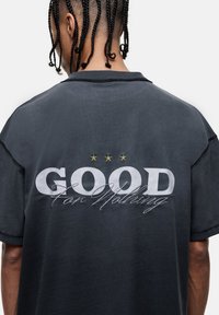 Czarna szaro-bawełniana luźna koszulka z wyraźnym białym napisem "DOBRZE" i "Za Nic" oraz trzema żółtymi gwiazdkami powyżej. Luźny krój.