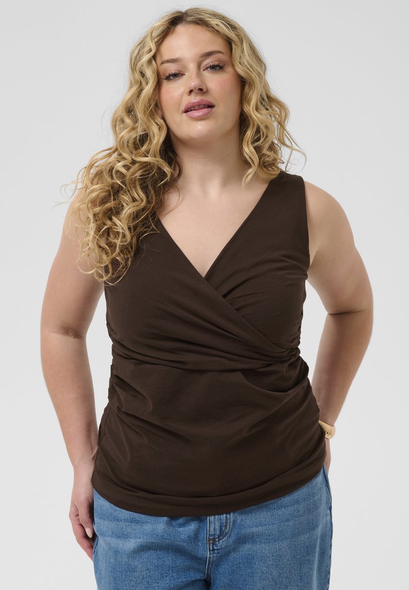 Femme aux cheveux bouclés portant un haut cache-cœur marron foncé sans manches et un jean bleu, debout les mains dans le dos.