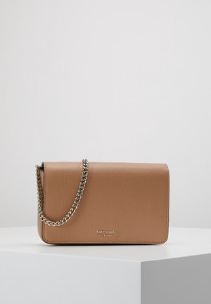 Sac à bandoulière en cuir marron Kate Spade avec bandoulière chaîne argentée sur surface d’exposition blanche sur fond gris.