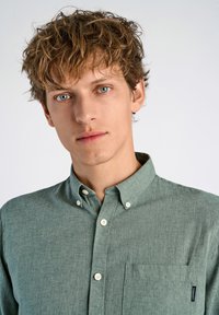 Camisa de botones verde claro con una tela texturizada, cuello y un único bolsillo en el pecho que presenta una pequeña etiqueta en el bolsillo.