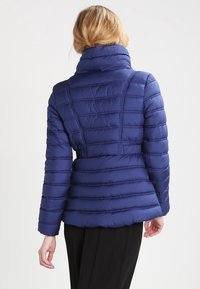 Tmavě modrá prošívaná bunda s vysokým límcem, prošívanou strukturou a přiléhavým pasem. Na zádech má vodorovné švy.
