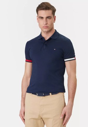 Giovane uomo che indossa una polo blu navy con polsini a righe rosse e bianche e pantaloni beige su sfondo bianco semplice.