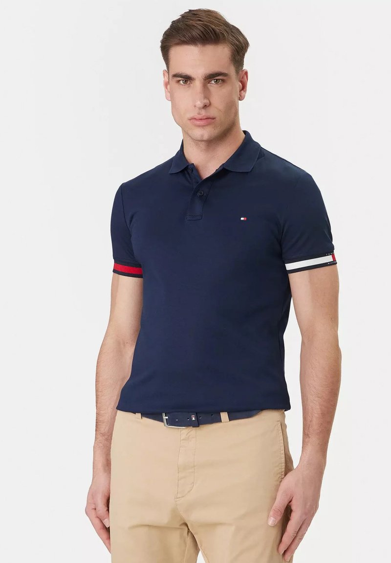 Jeune homme portant un polo bleu marine avec des poignets de manches rayés rouges et blancs, ainsi qu'un pantalon beige, sur un fond blanc uni.