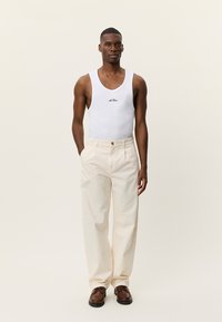 Λευκό αμάνικο τοπ με λογότυπο, εκρού παντελόνι με φαρδιά μπατζάκια και καφέ παπούτσια. Το σύνολο χαρακτηρίζεται από απλές γραμμές και ουδέτερους τόνους.