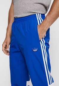 Blå idrottsbyxor i lätt tyg, med vita sidostråk och en broderad logotyp på vänster lår. Elastisk midja.