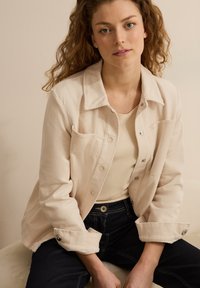Ljust beige kordjacka med krage, knappstängning och uppvikta ärmar, som bärs över en åtsittande krämfärgad skjorta och mörka jeans.