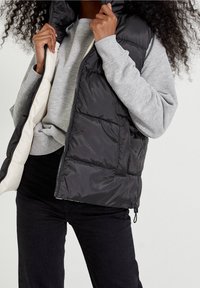 Czarna kamizelka puchowa z wysokim kołnierzem, z dwukolorowym wnętrzem i przeszywanymi panelami. Nosi się ją na szarym swetrze. Zamek błyskawiczny z przodu.