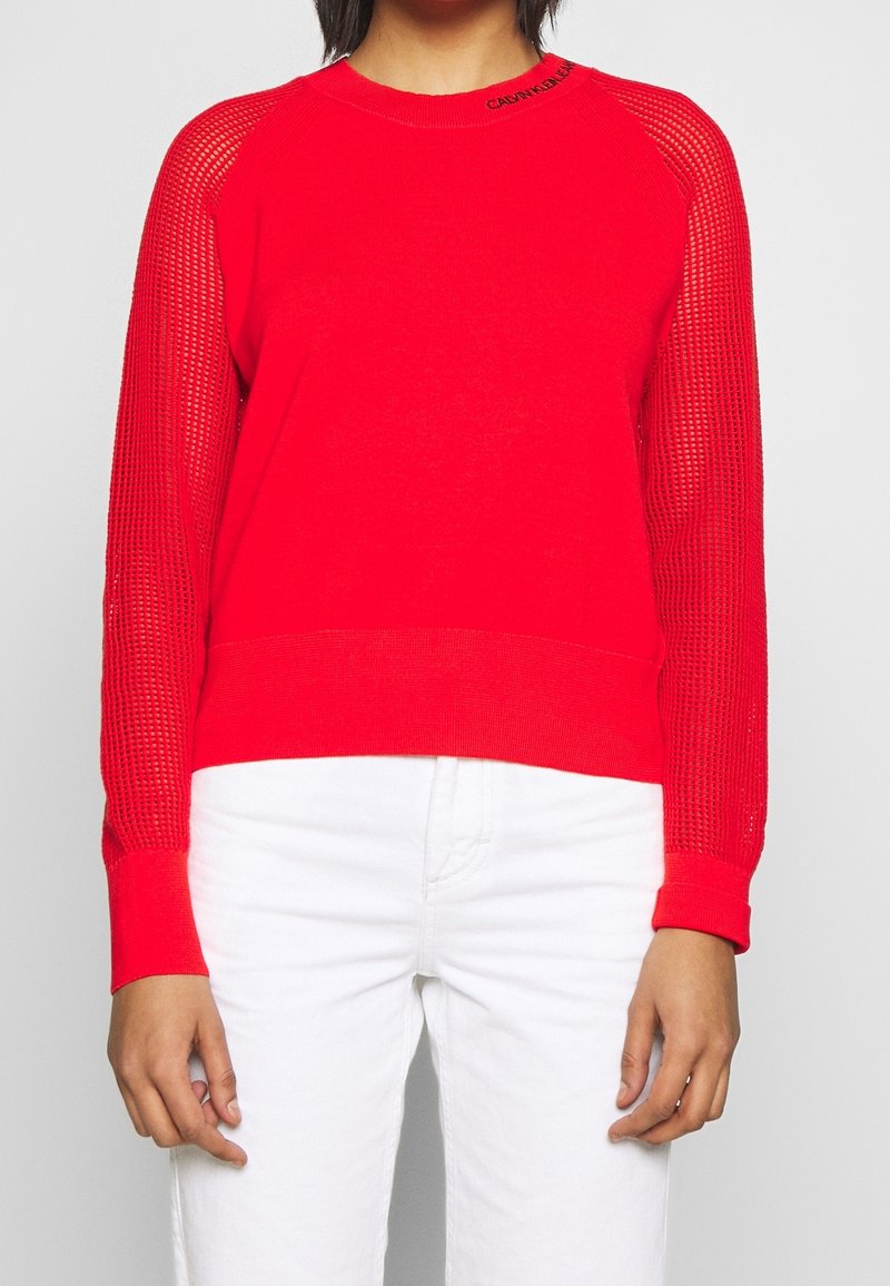 Personne portant un pull rouge vif avec des manches en résille et un pantalon blanc, moitié inférieure visible, sur fond gris uni.