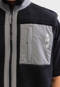 Gilet sans manches en polaire noire avec un devant en nylon gris. Comprend une poche zippée et un logo imprimé sur la poitrine. Tissu texturé.