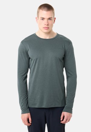 Jonge man met kort haar, gekleed in een nauwsluitend grijs shirt met lange mouwen en donkere broek, staand tegen een effen lichte achtergrond.