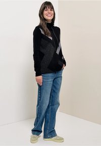 Zwart vest met ritssluiting en grijze geometrische patronen, gecombineerd met lichtblauwe jeans met wijde pijpen en beige sneakers. Effen achtergrond.