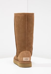 Stivale marrone in suede alto fino al ginocchio con finitura texturizzata, caratterizzato da cuciture a vista e suola in gomma color talpa. Il logo UGG è visibile sul tallone.