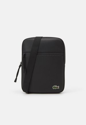 Svart slank crossbody-veske laget av teksturert materiale, med en frontlomme med glidelås, justerbar stropp, og grønn krokodillelogo.